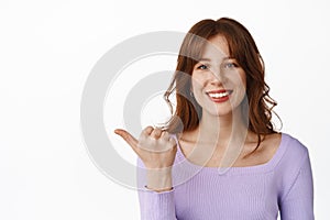 Close up of pretty girl with bangs, new hairstyle and make up on face, pointing thumb left, smiling happy with white