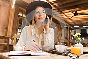 Close up of a pretty girl in hat sitting at the cafe table indoors, talking on mobile phone, taking notes in a notebook,