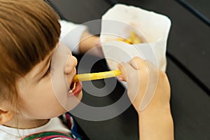 Close-up of a pretty little girl eating delicious French fries while sitting at a table in a fast food cafe. Cute baby girl eats u