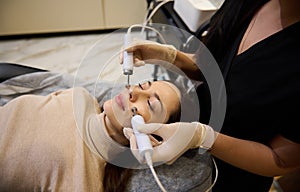 Close-up pretty woman receives facial microcurrent treatment from beautician at spa salon. Cosmetologist using electrical impulses