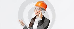 Close-up of professional smiling asian female entrepreneur at factory, chief engineer in safety helmet and suit, talking