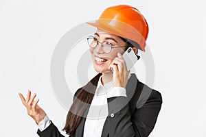 Close-up of professional smiling asian female entrepreneur at factory, chief engineer in safety helmet and suit, talking