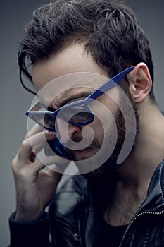 Close up of a profile a frowning bearded man in sunglasses, talking on a mobile phone, isolated on a grey background.