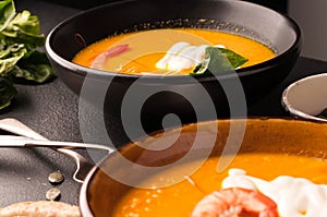 Close up pumpkin soup with shrimps, pumpkin seeds in dark bowls and bread, silver spoons