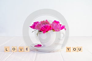 Close up purple pink peony flowers bouquet in a decorative cup and saucer and message I love you spelled in wooden blocks on white