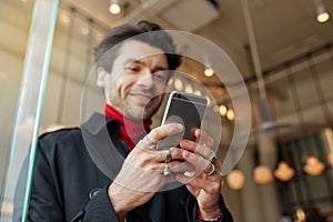 Close-up of raised man`s hands with rings keeping mobile phone while posing over city cafe background, texting message to friends