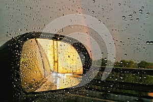Close-up of rear view car mirror with sunset and rainbow and raindrops