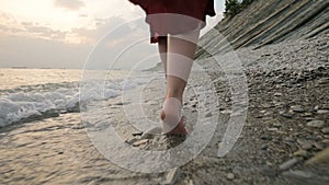 Close-up rear view of legs Caucasian girl walks along the water on the stone coast of the sea towards the setting sun at