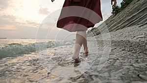 Close-up rear view of legs Caucasian girl walks along the water on the stone coast of the sea towards the setting sun at