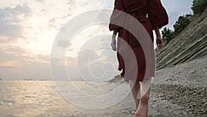 Close-up rear view of legs Caucasian girl walks along the water on the stone coast of the sea towards the setting sun at