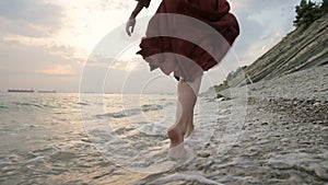 Close-up rear view of legs Caucasian girl walks along the water on the stone coast of the sea towards the setting sun at