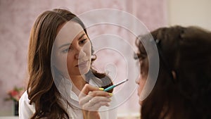 Close-up rear view of working professional makeup artist applying eyeshadow to eyelids of young pretty woman in beauty