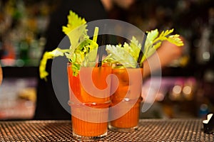 Close up of red bloody mary cocktails on bar top with green fresh celery leaves and bartender at night