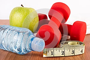 Close up of red dumbbell, water in bottle, green fresh apple with dew and measuring tape on wooden floor