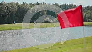 Close-up of red flag waving in wind on golf course with lake and two golfers against a background of green forest and
