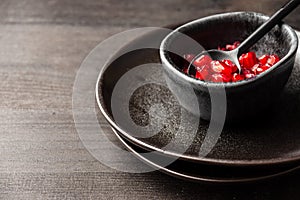Close-up of red pomegranate seeds in bowl with dark spoon and black dishes, on dark wooden table, horizontal,