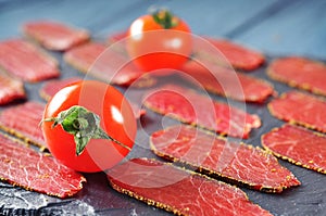 Close-up of red smoked meat slices with spices and ripe tomatoes on a dark cutting board with wooden blue table