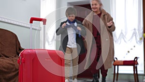 Close-up red suitcase with blurred woman and boy putting on clothes taking luggage leaving home for vacations. Caucasian