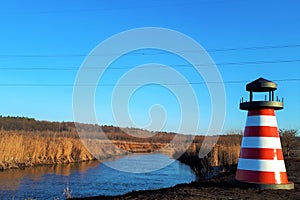 close-up of a red and white lighthouse near a river, against a background of reeds and a classically blue sky