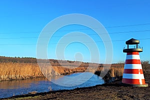 Close-up of a red and white lighthouse near a river, against a background of reeds and a classically blue sky