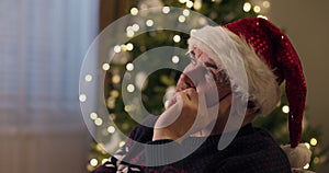 Close-up of retired man sitting in chair resting head on arm. He is wearing warm sweater and is wearing Santa hat. A man