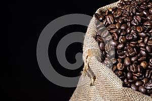 Close up roasted coffee beans in a burlap hessian sack on a wooden surface and isolated in black. Coffee beans scattered.