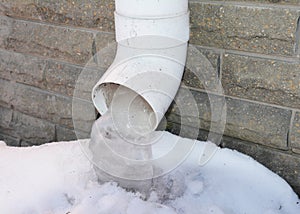 A close-up of a roof gutter downpipe, downspout with frozen water, icicles near the house foundation in winter