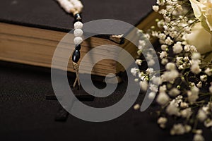 Close up of a rosary beads over a holy bible with blurred white small flowers, black background