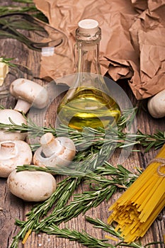 A close-up of rosemary twigs, yellow uncooked pasta, mushrooms, and olive oil. Fresh ingredients on a wooden table