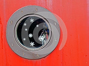 Close up of a round metal porthole on a red boat with snowflakes and glitter on the window and shiny charms hanging inside