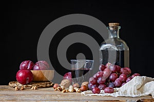 Close-up of rustic bottle and glass with red wine, grapes, plums and nuts on wooden table, black background