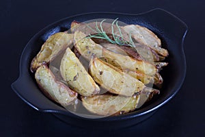 Close up rustic roasted potatoes with rosemary branch on black plate on black background