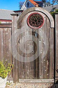 Close-up of rustic wooden gate with decorative round wrought iron window and a planting trowel for a knocker with two story house