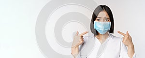 Close up of sad asian girl in medical face mask pointing at her head and looking upset, standing over white background