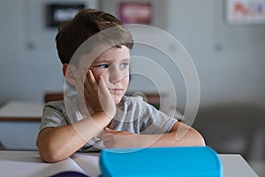 Close-up of sad caucasian elementary schoolboy with hand on chin sitting at desk in classroom