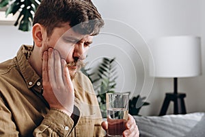 Close up of sad young man feel terrible toothache after drink cold water. Male 30s sitting on sofa at home touching cheek, feel