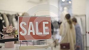 Close-up of Sale announcement in clothing store with blurred female shoppers choosing clothes at the background. Wealthy