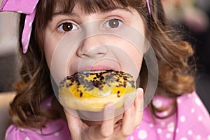 Close up school girl eating donut while sitting at table at home. Portrait of sweet girl choosing donut indoors. Funny