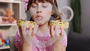 Close up school girl eating two donut while sitting at table at home. Portrait of sweet girl choosing between two donuts