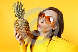 Close up selfie shot of amazed young woman in heart glasses holding fresh ripe pineapple fruit isolated on yellow orange