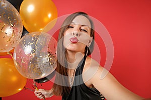 Close up selfie shot of woman in black dress celebrating blowing lips kiss hold air balloons isolated on red background