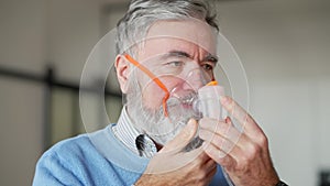 Close-up of senior bearded man putting on nebulizer indoors. Ill sad Caucasian retiree using inhaler for chronic