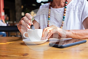 Close up on senior woman`s hand holding a white coffee cup while relaxing for a break at coffeeshop, Wooden table and mobile phon