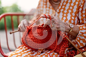 Close-up of senior woman sitting outdoor and knitting red scarf.