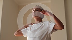 Close-up of serious little kid stretching neck for exercise during home workout standing in light living room. Pretty