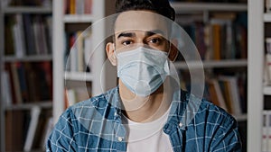 Close-up serious pensive young man sitting indoors alone wearing protective surgical mask hispanic guy student looking
