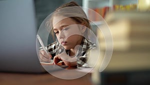 Close-up of serious primary child school girl doing homework writing notes in paper notebook and typing on laptop.