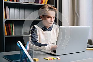 Close-up of serious pupil boy using typing laptop computer doing online lesson via Internet at desk, selective focus.