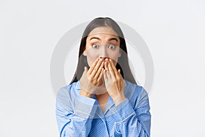 Close-up of shocked and astounded cute asian girl in blue pajama realise something, holding hands on mouth and pop eyes