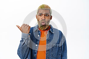 Close up of shocked Black man pointing left, looking concerned and startled, stands over white background